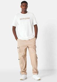 Lichtbeige cargo-broek met meerdere zakken, gecombineerd met een witte t-shirt met een beige grafisch logo, en witte sneakers.