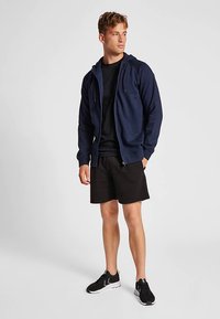 Navyblauwe capuchontrui met rits en trekkoorden, gedragen over een zwart T-shirt en gecombineerd met zwarte korte broek. Model draagt zwarte sportschoenen.