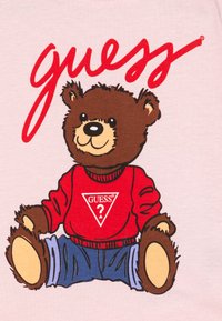 T-shirt rose clair avec un ours en dessin animé à la fourrure brune, portant un pull rouge et un jean bleu, accompagné du logo "guess" en rouge et en écriture cursive.