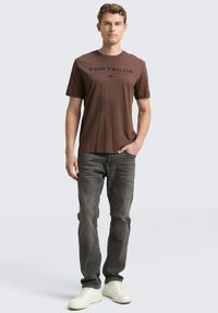 Bruine katoenen t-shirt met een ronde hals en een donkere print met de tekst "TOM TAILOR" op de voorkant, gecombineerd met grijze jeans en witte sneakers.