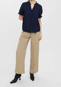 Blouse bleu marine à manches courtes avec un décolleté en V et une fermeture à boutons ; associée à un pantalon beige à jambes larges et des bottines à talons noires.
