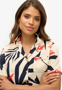 Camisa de manga corta en color crema con estampado de hojas en azul marino y rojo, con cuello, un bolsillo en el pecho y un acento de logo prominente.
