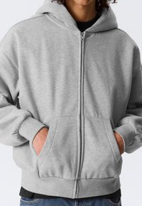 Personne portant un sweat à capuche gris clair avec une fermeture éclair, les mains dans les poches avant, et un t-shirt noir en dessous, sur un fond blanc.