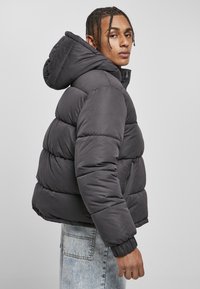 Grau gefütterte Jacke mit Kapuze, die über gesteppten Abschnitten, elastischen Bündchen und einer Reißverschluss-Seitentasche verfügt. Über hellblauen Jeans getragen.