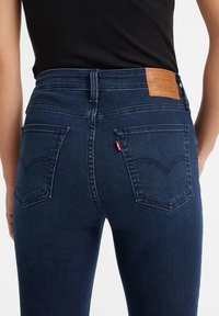 Donkerblauwe jeans met een hoge taille, twee achterzakken en een bruine leren label. De stof heeft een gladde textuur met subtiele stikseldetails.