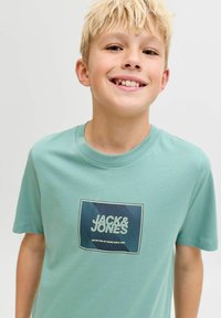 Licht turquoise katoenen T-shirt met een rechthoekige grafische print waarop "JACK & JONES" staat in vetgedrukte witte en blauwe letters. Ronde halslijn, korte mouwen.