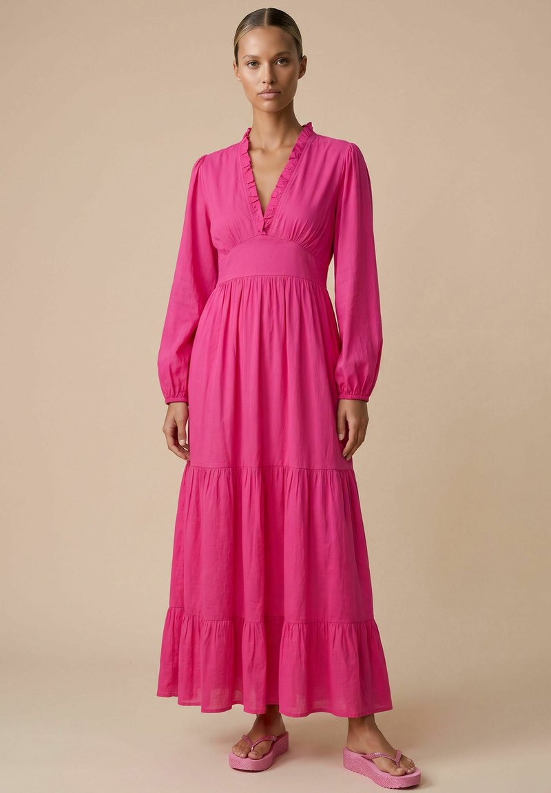 Femme aux cheveux tirés en arrière, portant une robe longue rose vif à manches longues avec un col à volants et une jupe à volants sur fond beige.