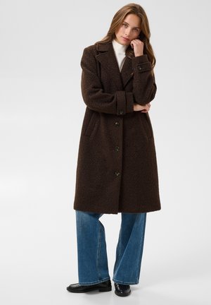 Jeune femme portant un long manteau texturé marron foncé, un jean bleu et des chaussures noires, debout avec la main près du visage sur un fond uni.