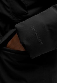 Czarna ręka o ciemnej karnacji w kieszeni czarnej kurtki Lululemon, pokazująca teksturowany materiał oraz logo Lululemon na rękawie.