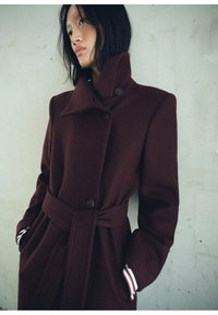 Manteau bordeaux avec un col montant, design double boutonnage, gros boutons et taille nouée. Présente des poignets rayés blancs. Texture douce.