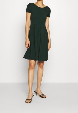 Dunkelgrünes Kleid mit kurzen Ärmeln, tailliertem Oberteil und schwingendem Rock. Kombiniert mit offenen Sandalen in Leopard-Print. Glatter Stoff.