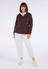 Felpa cappuccio marrone con scollo a V e stringhe, abbinata a pantaloni bianchi cropped. Il tessuto sembra morbido; il design complessivo è casual.