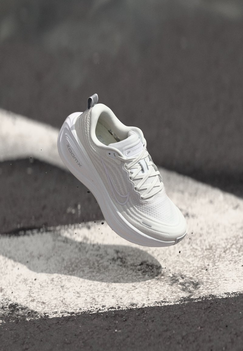 Chaussure de sport blanche avec lacets visibles et semelle "ZoomX", capturée en plein air au-dessus d'une surface d'asphalte noir et blanc.