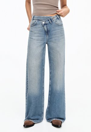 Personne portant un jean bleu délavé taille haute à jambes larges avec une fermeture à boutons asymétrique, associé à des chaussures marron pointues et un haut beige.