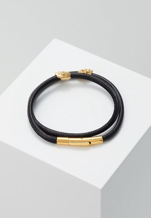 Zwarte dubbelgewonden leren armband met gouden schedelaccenten en een gouden magnetische sluiting, gepresenteerd op een wit voetstuk.