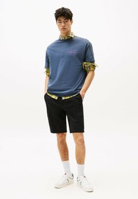 Camiseta de algodón navy con logo rosa, llevada sobre una camisa de cuadros amarilla. Combinada con pantalones cortos cargo negros y zapatillas blancas con detalles.