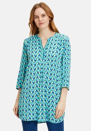 Turquoise tuniek met een V-hals en ¾ mouwen, met een herhalend golfpatroon in groene en blauwe tinten. Lichtgewicht stof.
