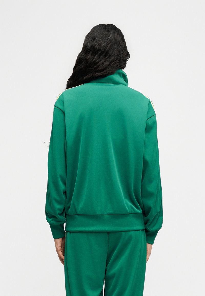 Person mit langen schwarzen Haaren, die einen grünen Trainingsanzug mit hohem Kragen trägt, von hinten vor hellem, einfarbigem Hintergrund.