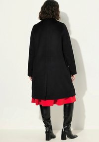 Cappotto nero lungo con taglio dritto, fessura posteriore e texture liscia, abbinato a una gonna rossa a strati e stivali neri sopra il ginocchio.