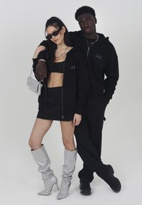 Black zip-up hoodie with purple lettering, paired with a black crop top and mini skirt. Model wears shiny silver knee-high boots. Black pants below.