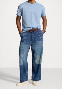 Man wearing a light blue Polo Ralph Lauren t-shirt, patchwork blue jeans with a brown belt, and white sneakers, hands in pockets.