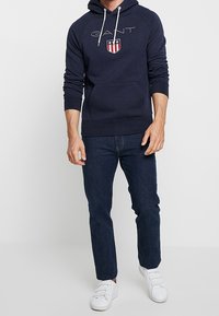 Man som bär en marinblå GANT-hoodie med sköldlogotyp, mörkblå jeans och vita sneakers, stående mot en enfärgad vit bakgrund.