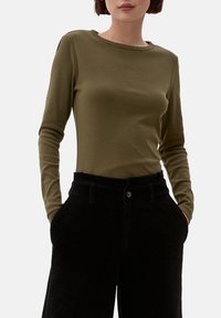 Maglietta a maniche lunghe verde oliva con scollatura tonda, abbinata a pantaloni in corduroy neri con tasche anteriori e chiusura a bottone.