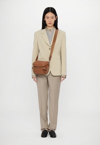 Brown leather crossbody bag with a top flap, chain accent, and a long strap, paired with a beige suit and gray shirt.