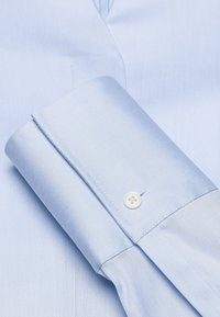 Camicetta azzurro chiaro con una texture liscia, dotata di chiusura a bottone sui polsini e di un design del colletto classico.