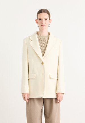 Blazer a spina di pesce crema con bavero a scollo, chiusura con bottone singolo e due tasche frontali, indossato sopra un top in maglia e pantaloni beige.