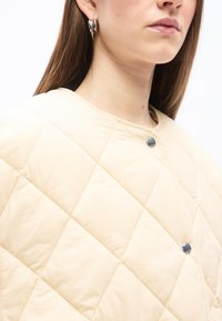 Γυναίκα με μπεζ μπουφάν καπιτονέ με ασημένια κουμπιά και ασημένια κρίκους σκουλαρίκια, εμφανιζόμενη από τον λαιμό μέχρι τον ώμο.