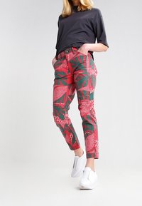 Rode spijkerbroek met groene bladmotieven en abstracte ontwerpen; gecombineerd met een zwarte top met korte mouwen en witte sneakers.