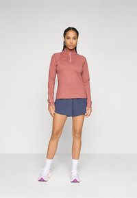 Langärmliges, rosafarbenes Sportoberteil mit halbem Reißverschluss, kombiniert mit marineblauen Shorts. Das Modell trägt weiße Socken und Sneakers mit rosa Akzenten.