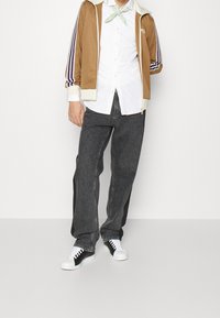 Brun sportjacka med vita och lila ränder, vit skjorta, svarta denimjeans och svartvita sneakers. Grön halsduk.