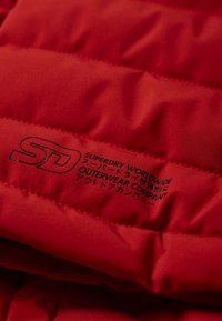 Czerwona pikowana kurtka z widocznymi szwami. Zawiera czarny nadrukowany tekst logo "SD Superdry Worldwide Outerwear Company." Miękka tekstura.