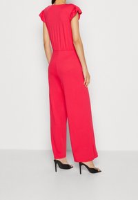 Red jumpsuit with cap sleeves, elastic waist, and wide leg design. Smooth texture and solid color, paired with black high-heel sandals.