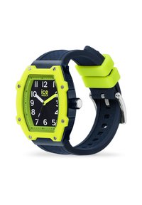 Reloj cuadrado con una cara negra, números blancos y un bisel amarillo brillante. Presenta una correa de goma azul marino con acentos texturizados.