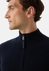 Mężczyzna w granatowym swetrze z ribbed, z pół zamkiem błyskawicznym i czarnym uchwytem oraz srebrnym detalem w kształcie litery "B", zwrócony w lewo na białym tle.