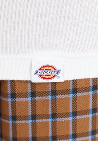 Weißer gerippter Stoff mit einem Etikett, das das "Dickies"-Logo zeigt, kombiniert mit einem braunen und blauen karierten Muster auf einer weichen Textiloberfläche.