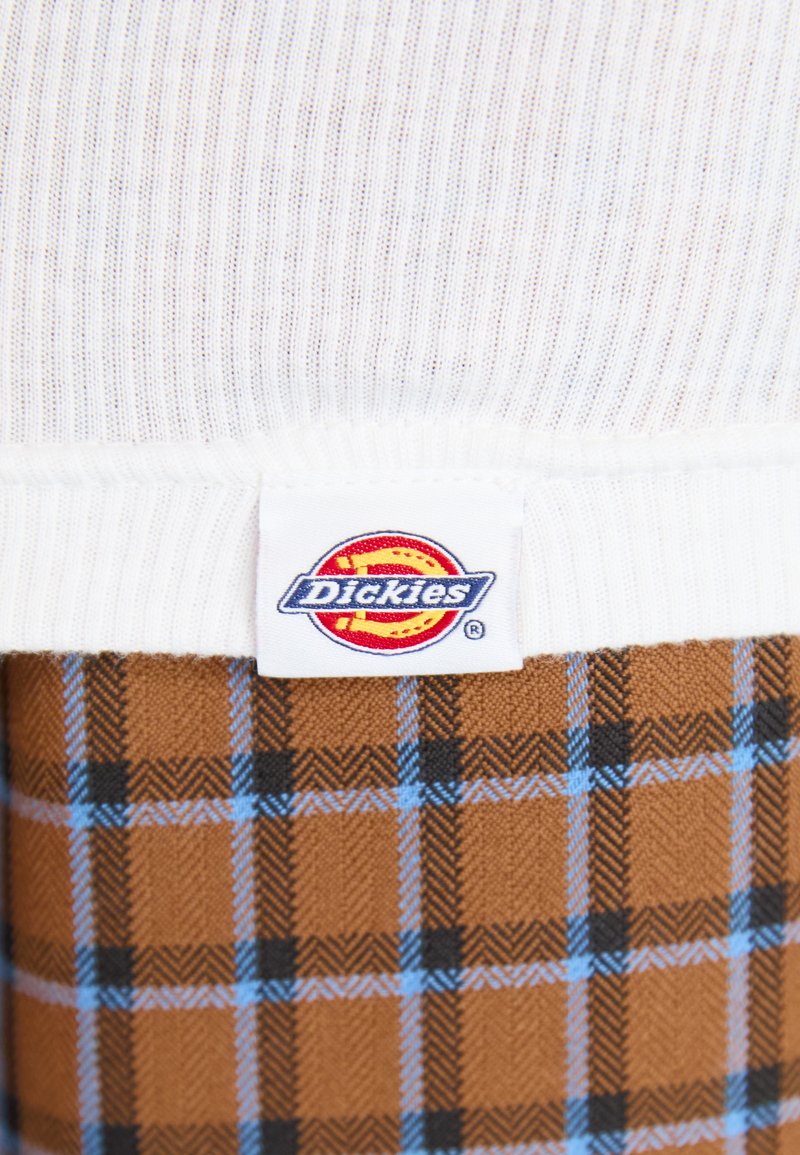 Weißer gerippter Stoff mit einem Etikett, das das "Dickies"-Logo zeigt, kombiniert mit einem braunen und blauen karierten Muster auf einer weichen Textiloberfläche.