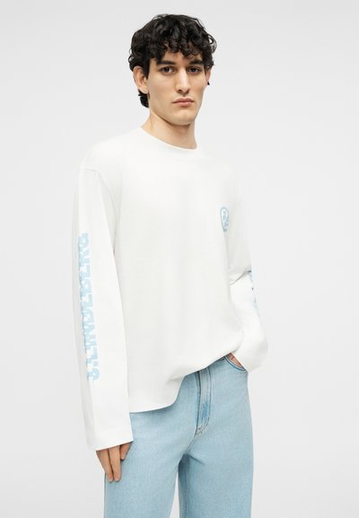 Νεαρός με σγουρά μαλλιά, φορώντας λευκό μακρυμάνικο μπλουζάκι με μπλε κείμενο στο μανίκι και ανοιχτόχρωμα τζιν, σε απλό φόντο.