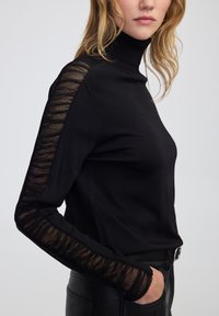 Vrouw in een zwart coltrui met doorschijnende, geplooide stoffen panelen langs de mouwen, gekleed in zwarte leren broek met hand in de zak.