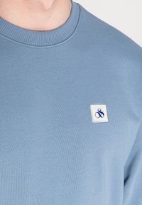 Niebieska bluza z miękkiego materiału, z prążkowanym dekoltem oraz małą białą metką z niebieskim napisem na klatce piersiowej.
