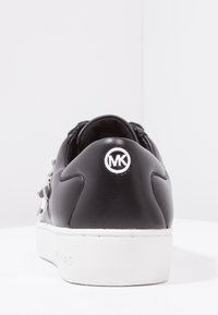 Black leather sneaker with a textured surface, featuring a white rubber sole and a circular logo on the heel. Accents include flat laces.