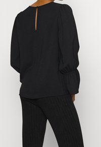 Blouse noire avec des manches bouffantes, un détail de dos en forme de clé et un tissu texturé. Associée à un pantalon à rayures sombres pour un look cohérent.