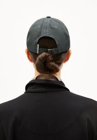 Cap in dark gray fabric with a curved brim and adjustable strap, worn backwards. Black jacket with a high collar and smooth texture.