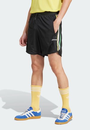 Person iført sorte mesh-shorts med grønne og gule striber, gule crew-sokker og blå Adidas-sneakers med hvide striber.
