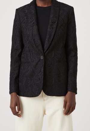 Zwarte blazer met kantstructuur en een knoopsluiting, gedragen over een zwart topje en crèmekleurige broek, getoond van schouders tot heupen.