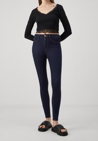 Mujer vistiendo jeans skinny azul oscuro, top corto negro de manga larga con borde de encaje y sandalias negras de plataforma, de pie sobre un fondo liso.