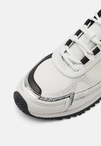Baskets blanches avec un matériau en maille texturée, des accents en cuir noir, des détails argentés et une semelle en caoutchouc épaisse dotée d'un motif de crampons.
